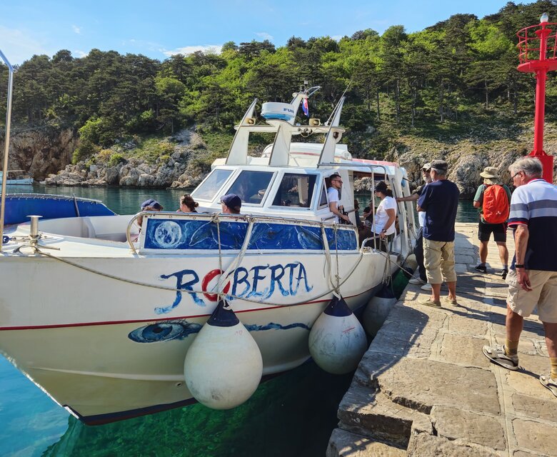 Ein Boot mit der Aufschrift "Roberta" liegt an einem Steg. Mehrere Personen steigen ein oder aus, während im Hintergrund grüne Hügel und klares Wasser zu sehen sind. Die Atmosphäre ist lebhaft und einladend.