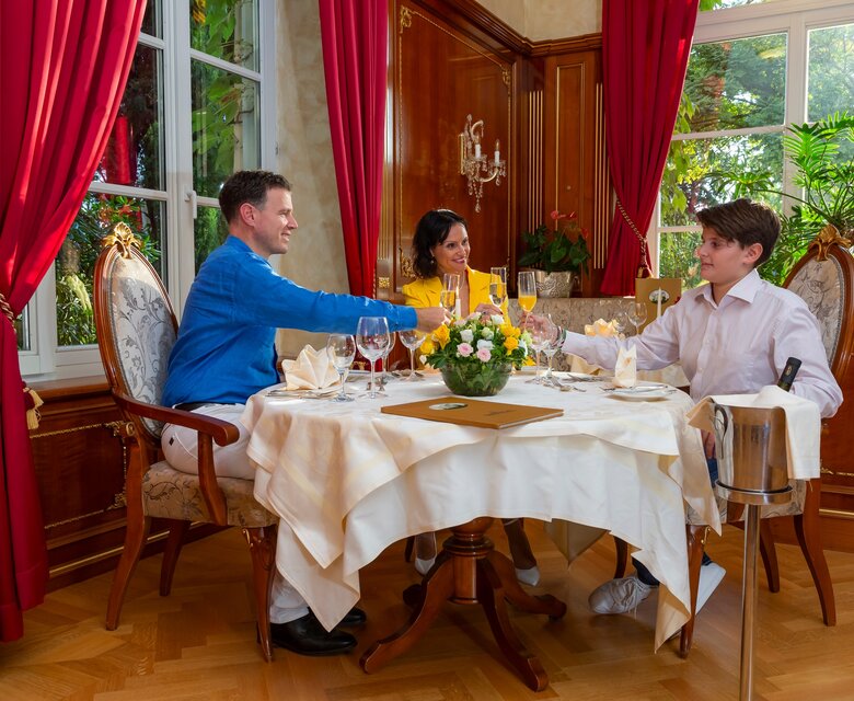 Ein elegantes Restaurant mit einem Tisch, der mit einer weißen Tischdecke und Blumen dekoriert ist. Drei Personen, zwei Männer und eine Frau, sitzen am Tisch und stoßen mit Gläsern an. Rote Vorhänge und Pflanzen im Hintergrund schaffen eine gemütliche Atmosphäre.