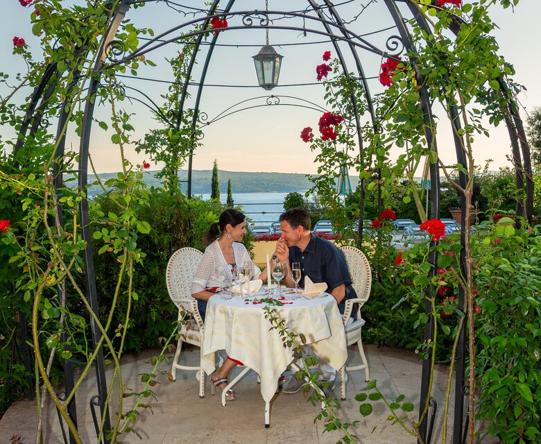 Ein romantisches Dinner für zwei in einem blühenden Pavillon, umgeben von roten Rosen. Das Paar sitzt an einem schön gedeckten Tisch mit Blick auf einen See und die sanfte Abenddämmerung.