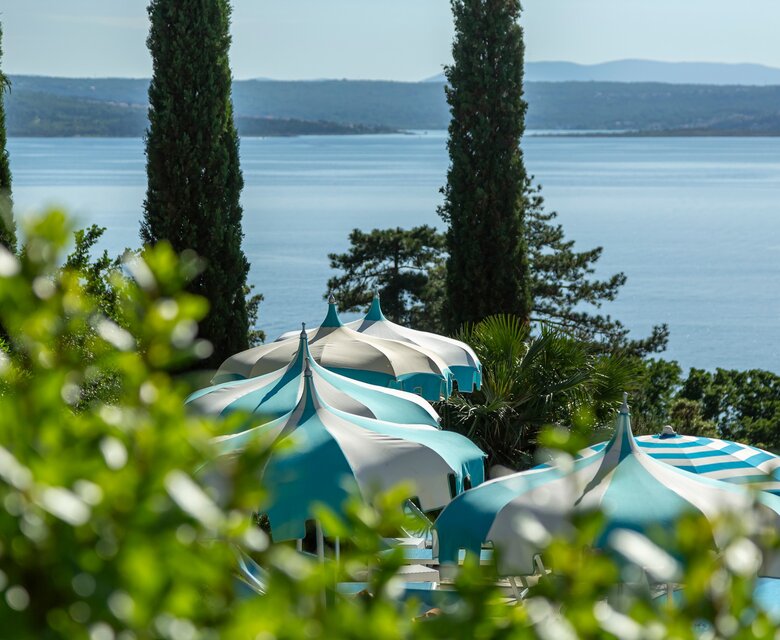 Blick auf eine ruhige Küstenlandschaft mit blauen und weißen Sonnenschirmen, umgeben von grünen Bäumen und Sträuchern. Im Hintergrund erstreckt sich der glitzernde See unter einem klaren Himmel.