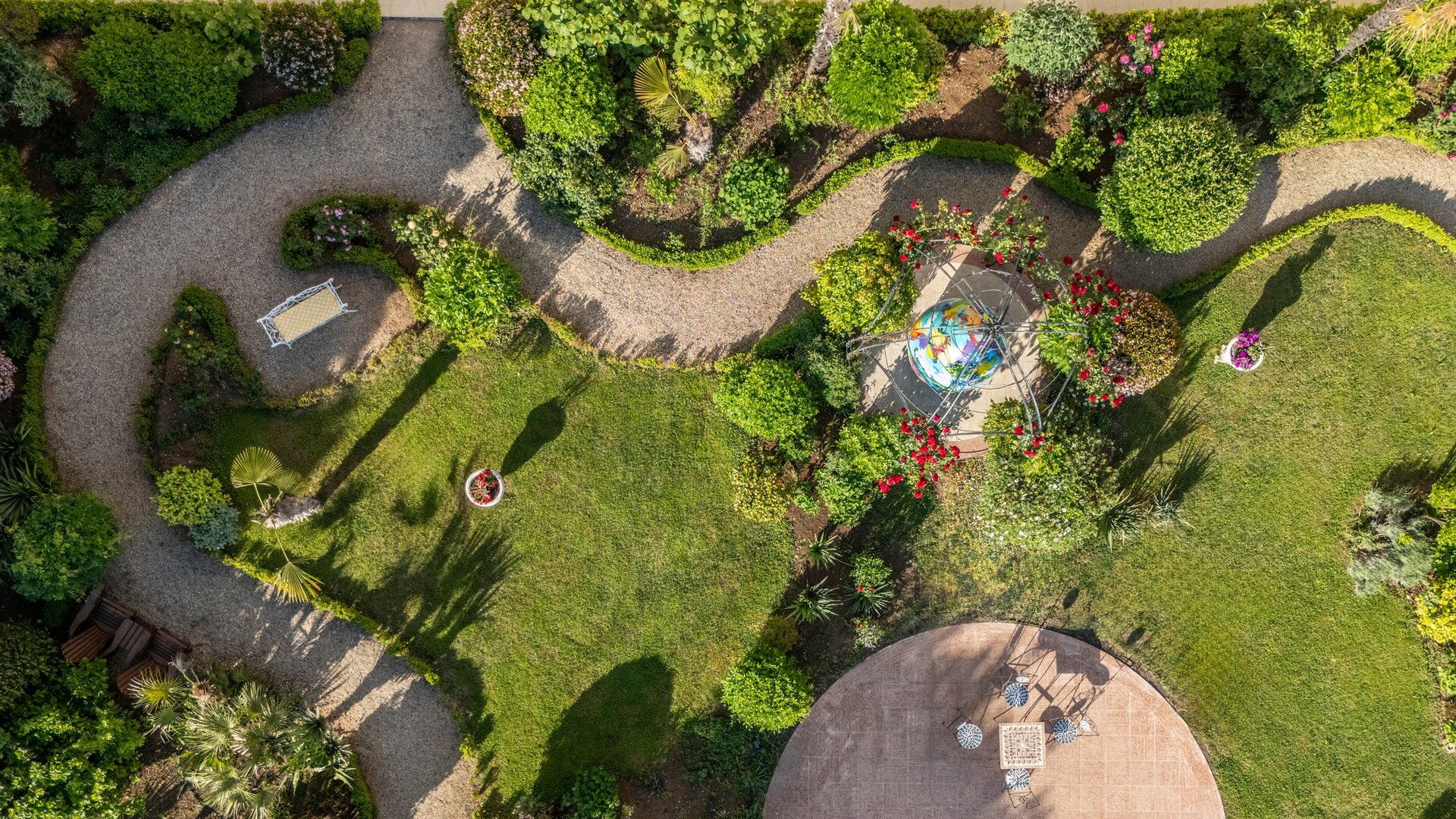 Luftaufnahme eines gepflegten Gartens mit geschwungenen Wegen, üppigem Grün und bunten Blumen. In der Mitte steht eine dekorative Gartenbank und ein runder Tisch mit Stühlen. Der Garten strahlt eine ruhige und einladende Atmosphäre aus.
