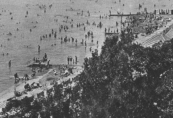 Ein belebter Strand mit vielen Menschen, die im Wasser schwimmen und am Ufer entspannen. Sonnenschirme und Liegen sind sichtbar, während einige Personen auf einem Steg stehen. Die Atmosphäre ist lebhaft und einladend, mit Blick auf das ruhige Wasser und den Himmel.