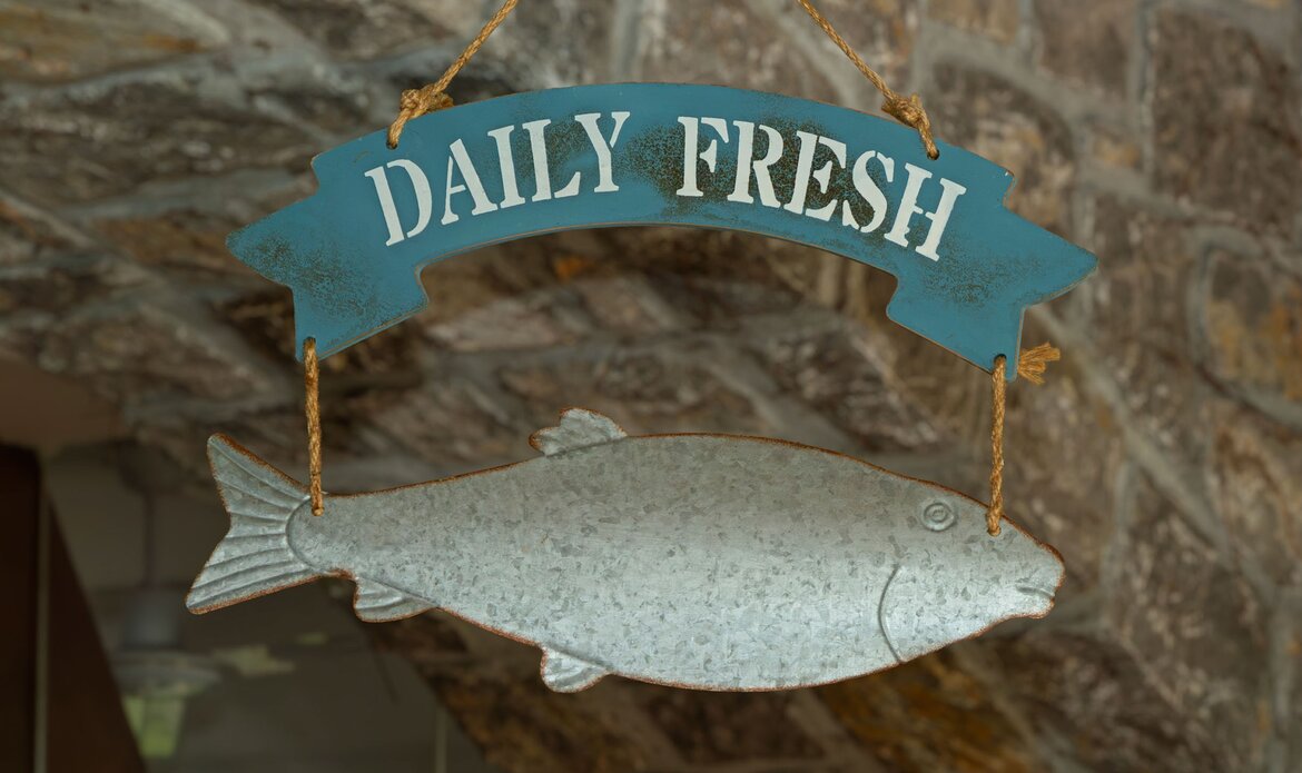 Ein hängendes Schild in Form eines Fisches mit der Aufschrift "DAILY FRESH" vor einer steinernen Wand. Das Schild vermittelt eine frische, maritime Atmosphäre.