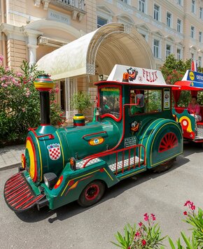 Bunter, nostalgischer Miniaturzug steht vor einem Hotel mit eleganter Fassade. Umgeben von blühenden Pflanzen und einem schattigen Vordach, strahlt die Szene eine einladende, fröhliche Atmosphäre aus.