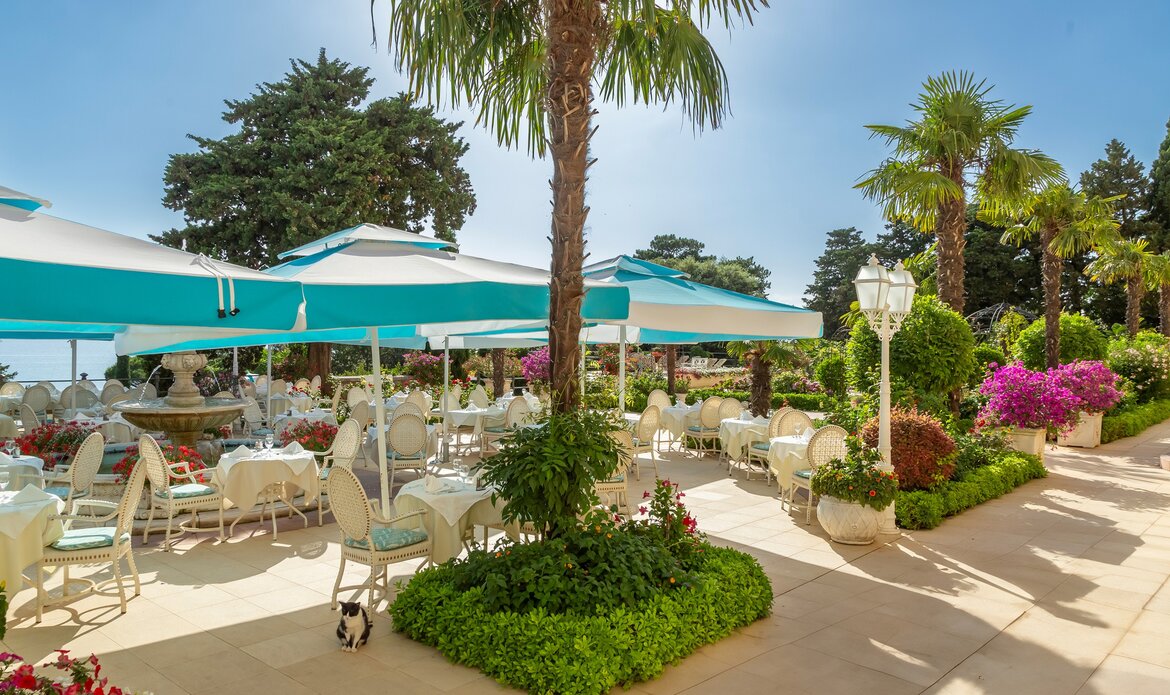 Ein einladender Außenbereich eines Restaurants mit weißen Tischen und Stühlen, umgeben von üppigen Pflanzen und bunten Blumen. Sonnenschirme bieten Schatten, während Palmen im Hintergrund für eine tropische Atmosphäre sorgen. Ein kleiner Hund läuft zwischen den Tischen.