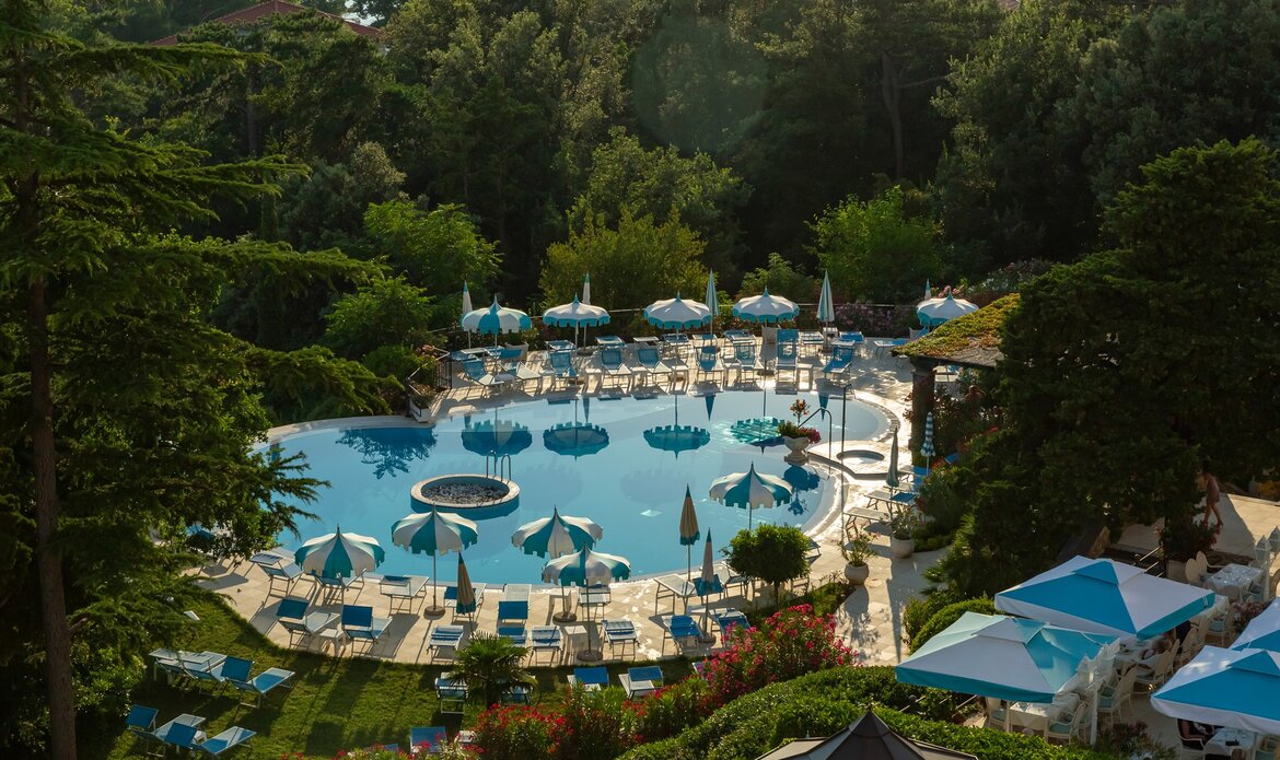 Ein weitläufiger Hotelpool umgeben von grünen Bäumen und bunten Liegen. Sonnenschirme spenden Schatten, während die Wasseroberfläche im Sonnenlicht glitzert. Eine entspannte Urlaubsatmosphäre mit vielen Liegen und einem einladenden Ambiente.