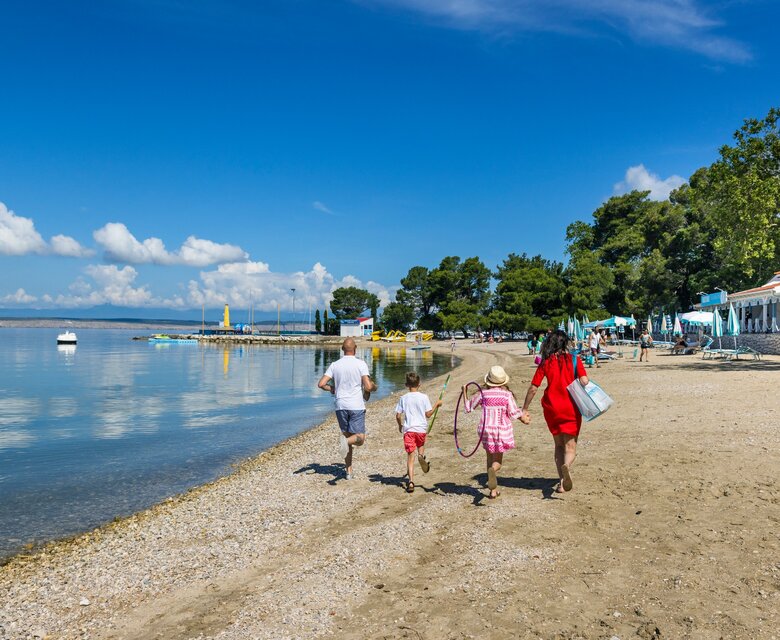 Eine Familie mit zwei Erwachsenen und zwei Kindern spaziert am Strand entlang, umgeben von ruhigem Wasser und Bäumen. Im Hintergrund sind Strandliegen und ein Restaurant sichtbar, während der Himmel strahlend blau ist.