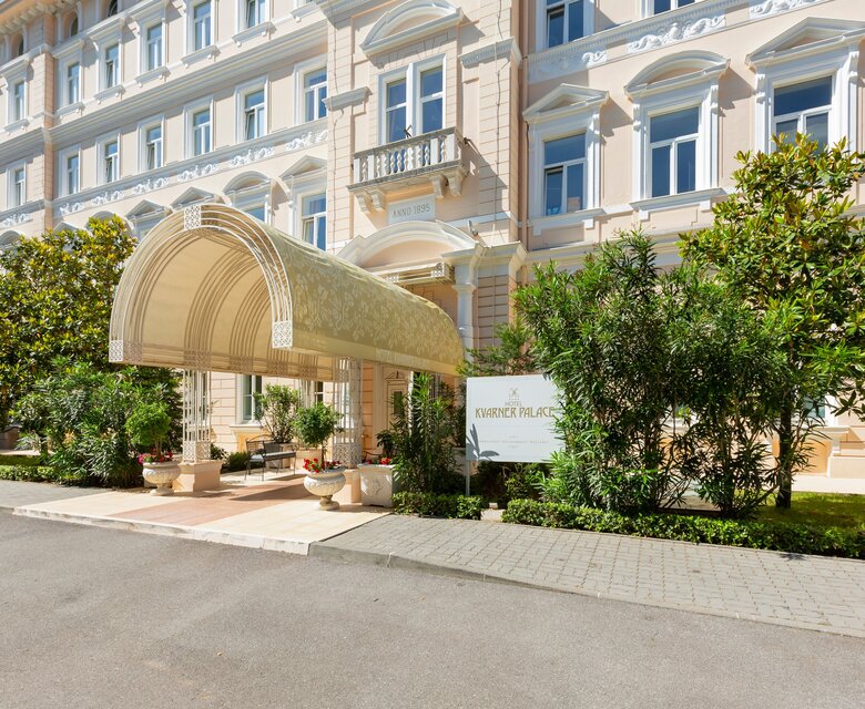 Eingang des Hotels mit einem eleganten Vordach, umgeben von grünen Pflanzen und einem gepflegten Außenbereich. Die Fassade des Gebäudes zeigt eine klassische Architektur mit großen Fenstern und dekorativen Elementen.