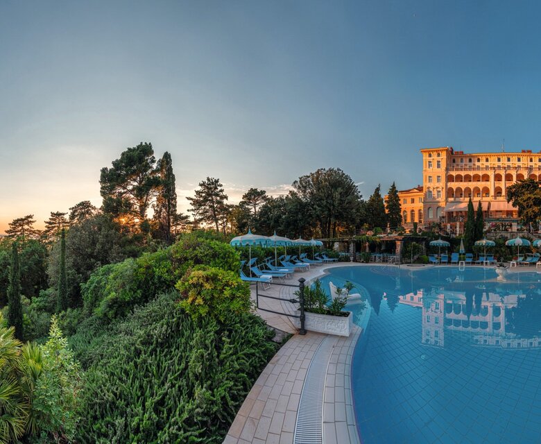 Ein Panoramablick auf einen luxuriösen Hotelpool, umgeben von üppigem Grün und Palmen. Im Hintergrund erhebt sich das elegante Hotelgebäude, während die Sonne am Horizont untergeht und eine entspannte Atmosphäre schafft.