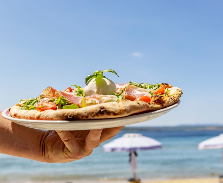 Eine Hand hält eine frisch zubereitete Pizza mit Tomaten, Schinken und einem Klecks Mozzarella. Im Hintergrund erstreckt sich ein schöner Strand mit Sonnenschirmen und dem glitzernden Wasser des Meeres unter einem klaren blauen Himmel.