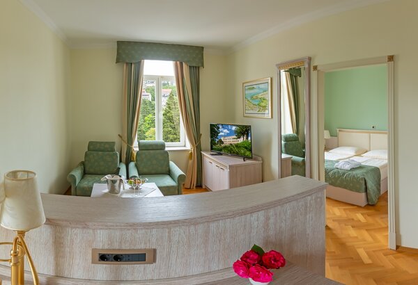Helles Hotelzimmer mit einem Wohnbereich, der zwei grüne Sessel und einen Fernseher umfasst. Im Hintergrund ist ein Schlafzimmer mit zwei Betten sichtbar. Große Fenster sorgen für viel Tageslicht u... Familienurlaub Kroatien ist so schön im Kvarner Palace