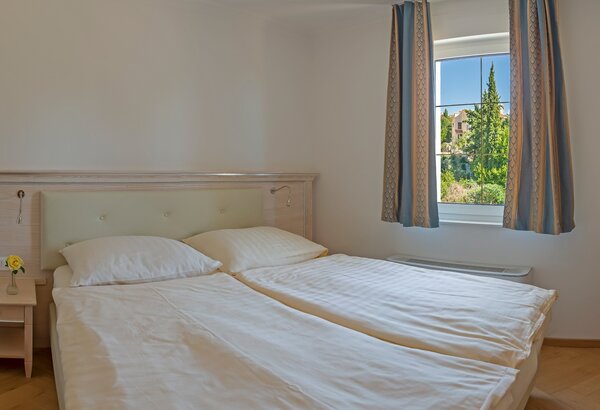 Helles Schlafzimmer mit zwei Einzelbetten, weißen Bettdecken und Kissen. Ein Nachttisch mit einer kleinen Blume steht neben einem Fenster, das Blick auf grüne Bäume bietet. Vorhänge in sanften Farb... Hotel mit Appartements für Familien, Kroatien - am Meer
