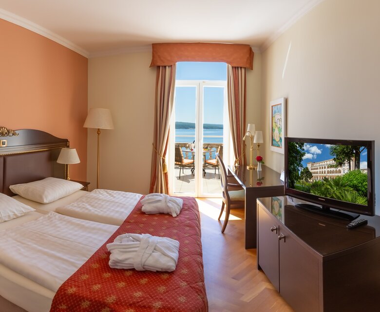 Ein helles Hotelzimmer mit zwei Einzelbetten, einem Fernseher und einem Schreibtisch. Große Fenster bieten einen Blick auf eine Terrasse mit Meerblick. Die Wände sind in warmen Farben gehalten, und die Einrichtung ist modern und einladend.