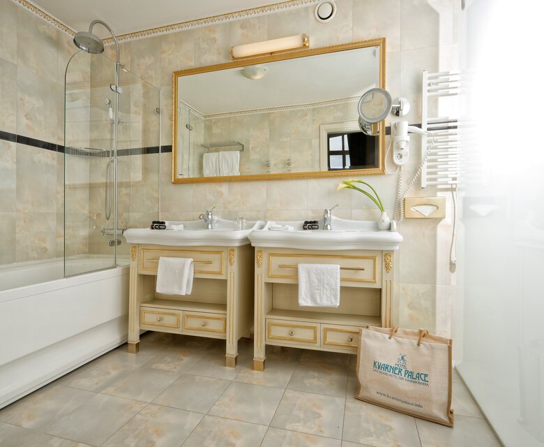Helles Badezimmer mit zwei Waschbecken, einem großen Spiegel und einer Dusche. Die Wände sind mit hellen Fliesen verkleidet, und eine Badewanne ist ebenfalls vorhanden. Ein Handtuchhalter und eine Pflanze ergänzen die Einrichtung.