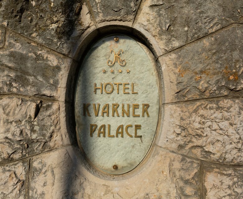 Schild des Hotels Kvarner Palace, eingelassen in eine steinerne Wand. Das Schild ist oval und zeigt den Hotelnamen in goldener Schrift auf hellem Hintergrund. Die umgebende Steinstruktur verleiht dem Eingang einen historischen Charme.