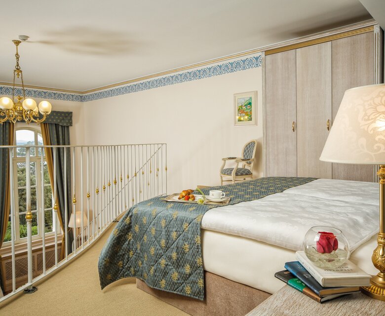 Ein gemütliches Hotelzimmer mit einem großen Bett, einer eleganten Bettdecke und einem Nachttisch mit einer Lampe. Im Hintergrund sind Fenster mit Vorhängen und ein kleiner Tisch mit Stühlen zu sehen. Eine Treppe führt zu einem oberen Bereich.