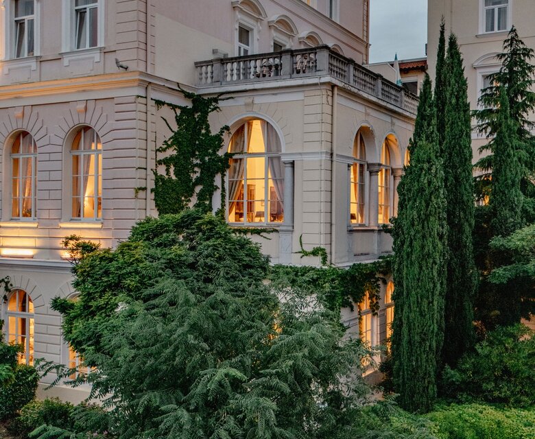 Eine elegante Hotelansicht mit einem historischen Gebäude, das von üppigem Grün umgeben ist. Die Fenster strahlen warmes Licht aus, während hohe Zypressen und Sträucher den Gartenbereich schmücken. Der Himmel ist leicht bewölkt, was eine ruhige Atmosphäre schafft.