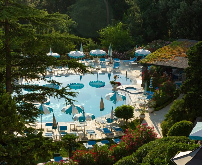 Ein einladender Poolbereich umgeben von üppigem Grün, mit blauen Liegen und Sonnenschirmen. Der Pool spiegelt das Sonnenlicht wider, während blühende Pflanzen eine entspannte Atmosphäre schaffen. Im Hintergrund ist eine gemütliche Bar zu sehen.