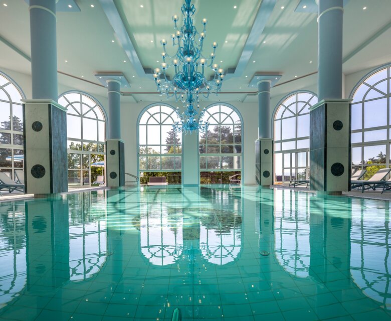 Eleganter Innenpool mit glitzerndem Wasser, umgeben von großen Fenstern, die viel Tageslicht hereinlassen. Ein stilvoller Kronleuchter hängt von der Decke, während Liegen am Rand des Pools stehen. Die Atmosphäre ist entspannend und einladend.