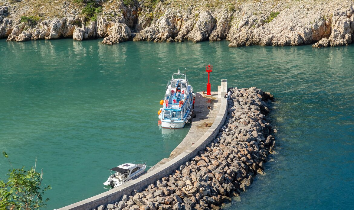 Ein Boot liegt an einem kleinen Hafen, umgeben von klarem, grünlichem Wasser. Der Hafen ist von einer geschwungenen Steinmauer umgeben, und im Hintergrund sind felsige Küstenlinien sichtbar. Ein rotes Signallicht steht am Ende des Stegs.