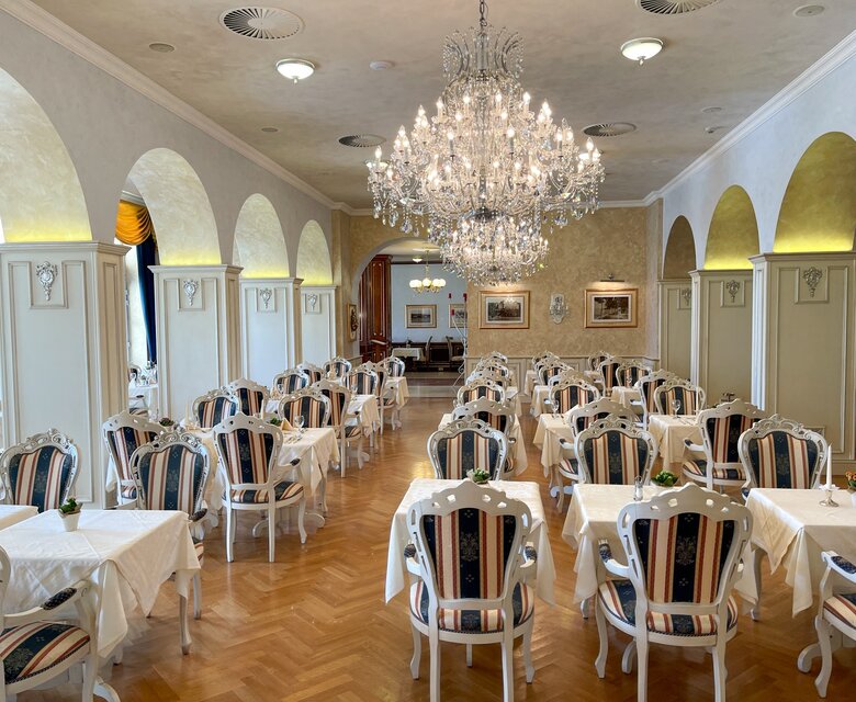 Elegantes Restaurant mit hellen Wänden und einem großen Kronleuchter. Die Tische sind mit weißen Tischdecken und gestreiften Stühlen dekoriert. Der Raum strahlt eine gehobene Atmosphäre aus, ideal für besondere Anlässe.
