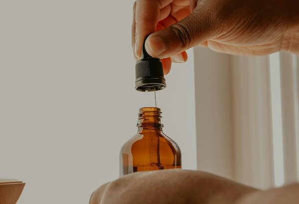 Eine Hand hält eine Pipette über eine braune Flasche, während sie einen Tropfen Flüssigkeit abgibt. Im Hintergrund ist ein heller Raum mit natürlichem Licht zu sehen.