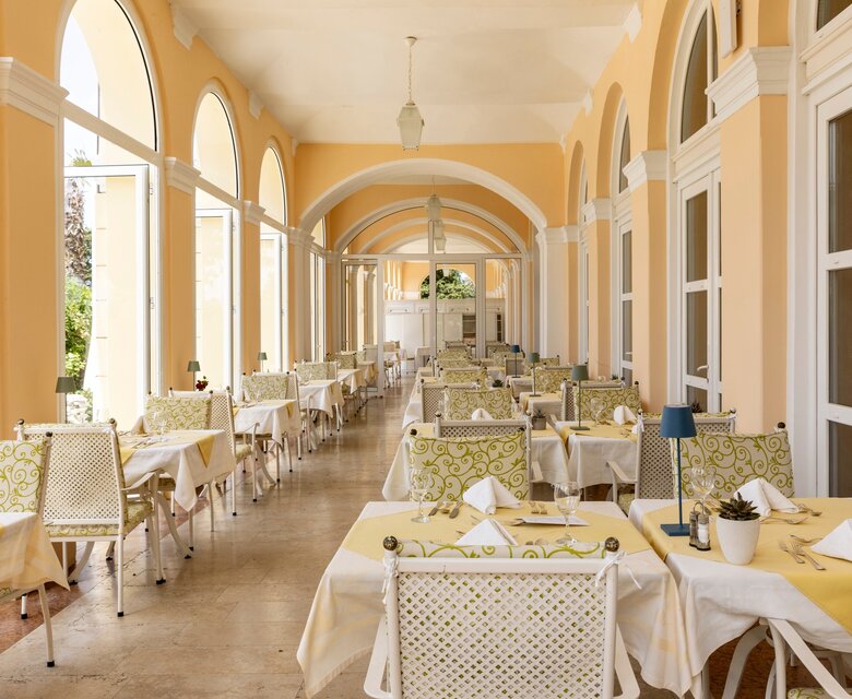 Ein eleganter Speisesaal mit weißen Tischen und Stühlen, die mit gelben Tischdecken und dekorativen Servietten gedeckt sind. Große Fenster mit Bögen lassen viel Licht herein und bieten einen Blick auf die Umgebung. Die Wände sind in sanften Gelbtönen gehalten, was eine einladende Atmosphäre schafft.