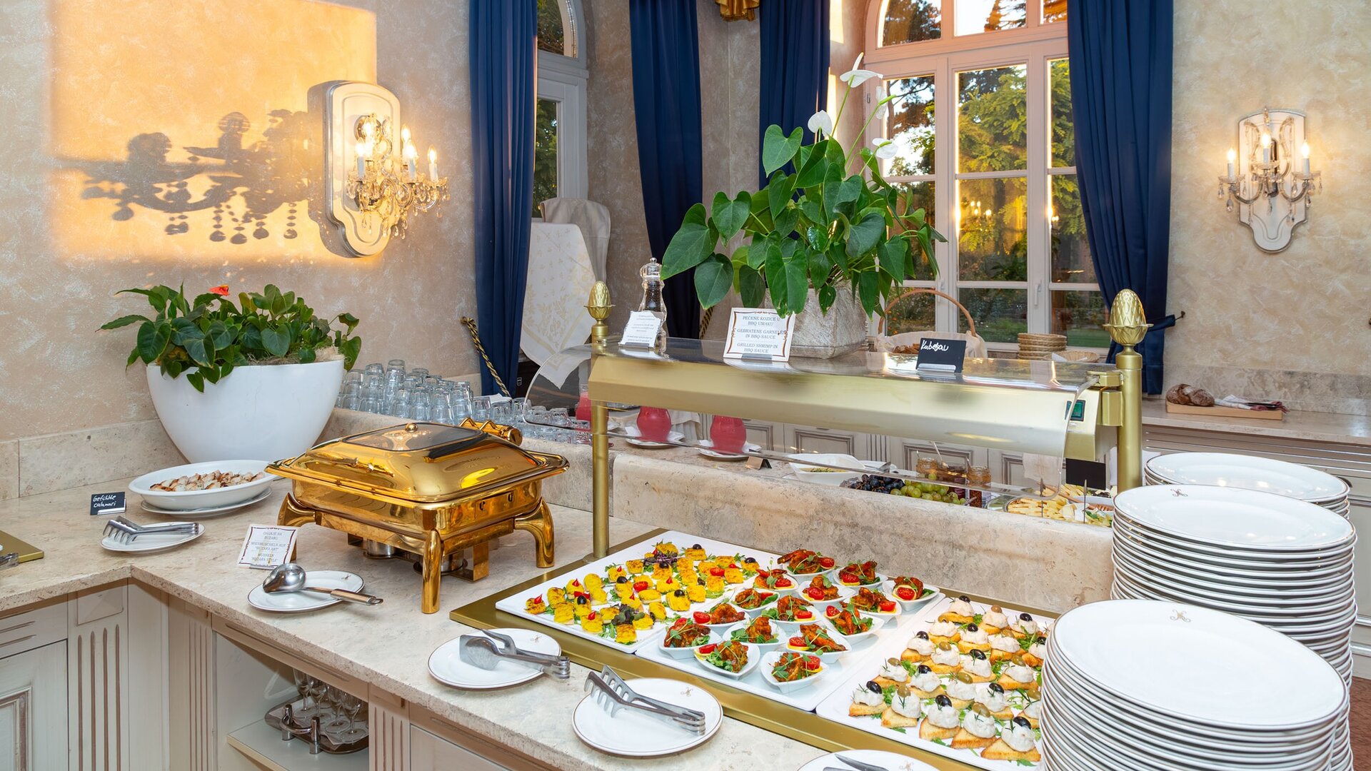 Ein eleganter Buffetbereich mit einer Auswahl an kunstvoll angerichteten Vorspeisen und Salaten. Im Hintergrund sind große Fenster mit blauen Vorhängen und Pflanzen zu sehen, die eine einladende Atmosphäre schaffen.