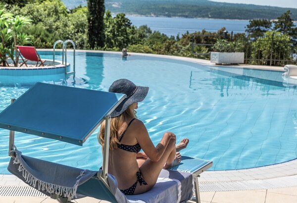 Eine Frau in einem Bikini sitzt entspannt auf einer Liege am Pool, umgeben von üppigem Grün und mit Blick auf das Wasser. Die Sonne scheint, und die Atmosphäre ist ruhig und einladend.