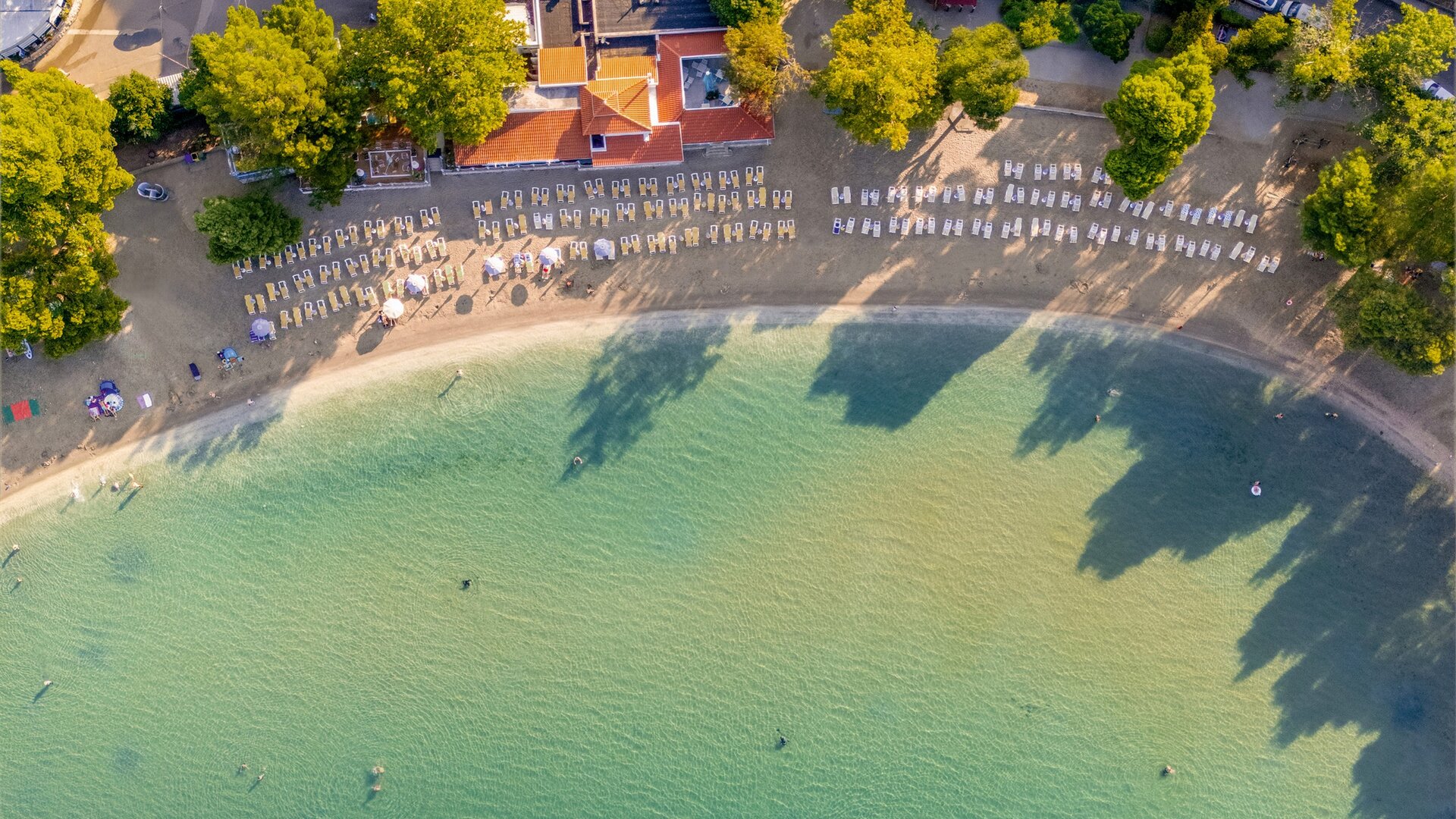 Luftaufnahme eines Strandes mit sanftem, klarem Wasser. Im Vordergrund sind Liegen auf dem Sand angeordnet, umgeben von Bäumen. Im Hintergrund sind einige Menschen im Wasser und am Strand zu sehen, was eine entspannte Urlaubsatmosphäre vermittelt.