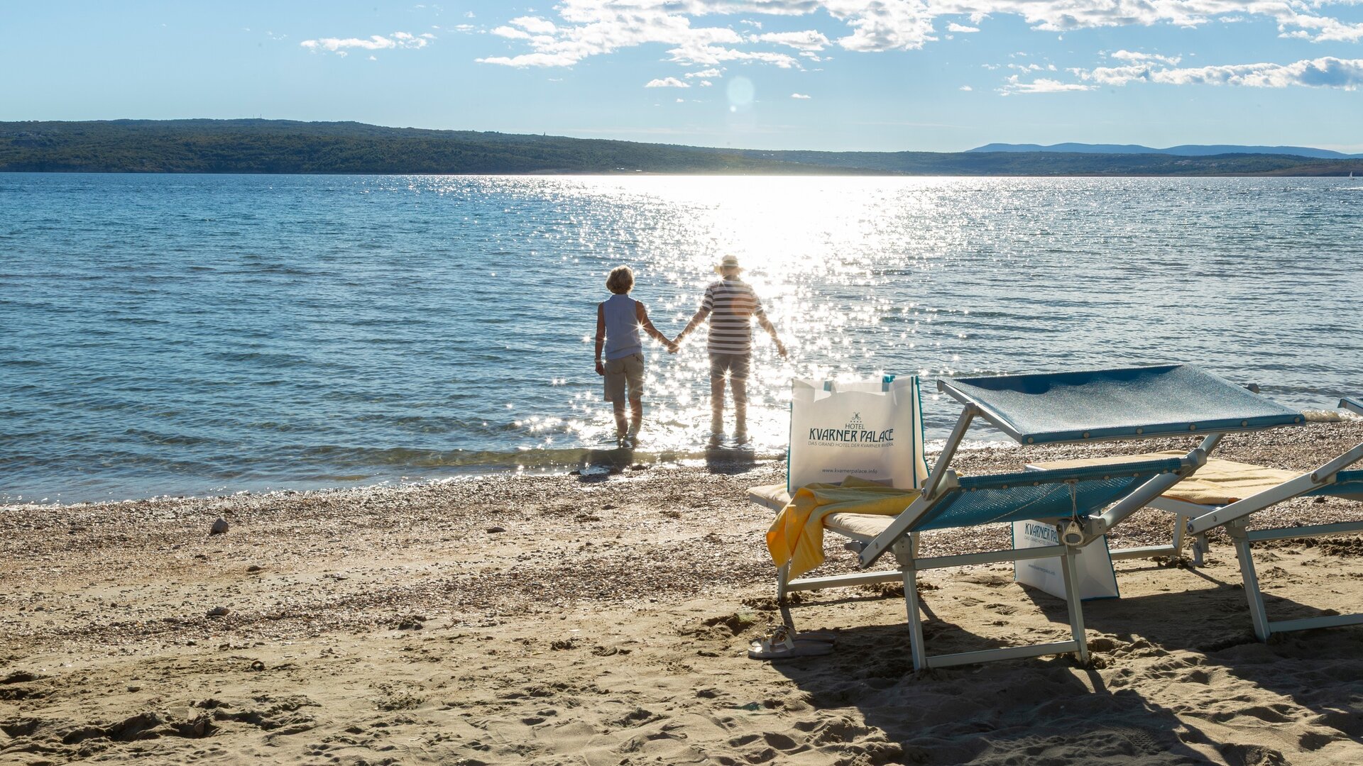 Ein Paar hält sich an der Hand und geht ins Wasser, während die Sonne auf die ruhige Wasseroberfläche scheint. Im Vordergrund stehen Liegen am Sandstrand. Die Landschaft ist friedlich und einladend.