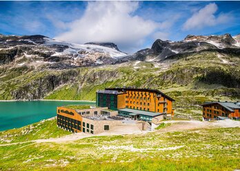 Ein modernes Hotel liegt malerisch am Ufer eines klaren Sees, umgeben von majestätischen Bergen und schneebedeckten Gipfeln. Die grüne Wiese im Vordergrund ergänzt die atemberaubende alpine Landschaft.