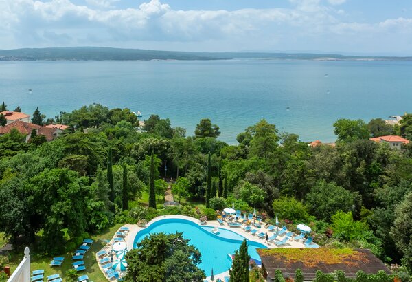 Blick auf einen blauen Pool umgeben von üppigem Grün und Sonnenliegen, mit dem ruhigen Wasser eines Sees im Hintergrund und sanften Hügeln am Horizont. Der Himmel ist teilweise bewölkt, was eine en... Meerblick Hotel Kroatien - Ihr Komfort Doppelzimmer