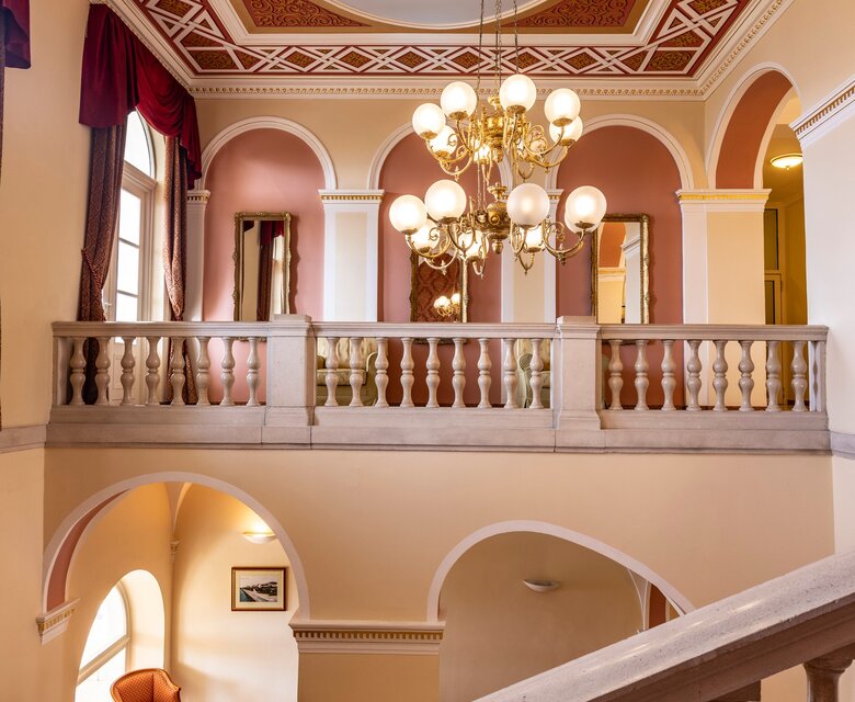 Ein eleganter Treppenaufgang in einem Hotel mit einem kunstvoll gestalteten Deckenmuster und einem großen Kronleuchter. Im Hintergrund sind gemütliche Sitzgelegenheiten und große Fenster, die viel Licht hereinlassen. Die Wände sind in sanften Farben gehalten.