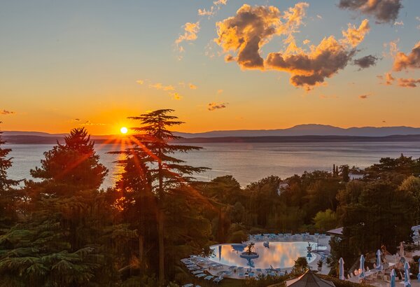 Sonnenuntergang über einem ruhigen See, umgeben von Bäumen und einem Poolbereich. Die warmen Farben des Himmels reflektieren sich im Wasser, während Sonnenstrahlen durch die Wolken scheinen. Eine e... Suite Crikvenica, Kroatien: Majestätisches Urlaubsgefühl