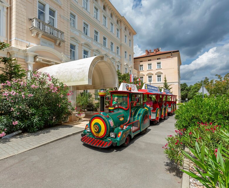 Bunte Mini-Eisenbahn vor einem eleganten Hotel, umgeben von blühenden Pflanzen und einem klaren Himmel. Die Atmosphäre ist einladend und freundlich, ideal für Familien und Touristen.