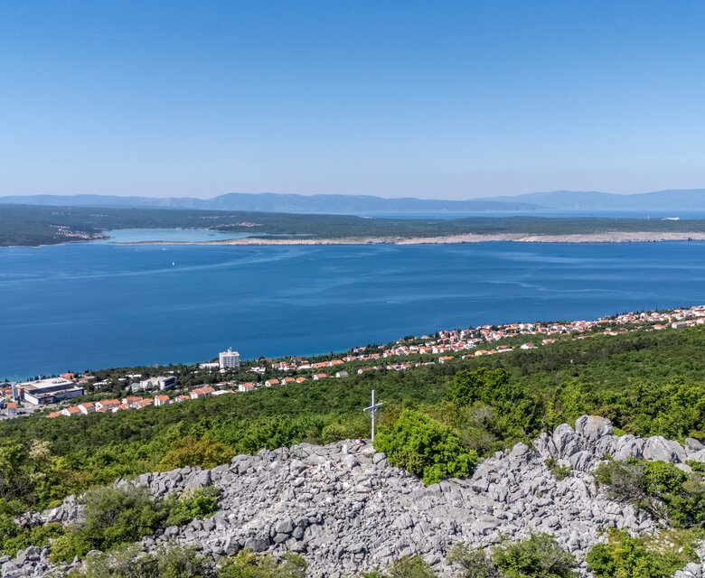Blick auf eine malerische Küstenlandschaft mit klarem, blauem Wasser und einer kleinen Stadt am Ufer. Im Hintergrund erstrecken sich sanfte Hügel und Berge unter einem strahlend blauen Himmel. Grüne Vegetation umgibt die felsige Vordergrundlandschaft.