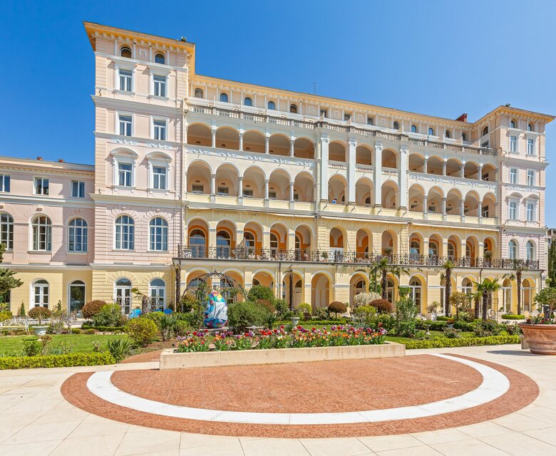 Ein elegantes Hotelgebäude mit mehreren Etagen, großen Bögen und Balkonen, umgeben von gepflegten Gärten und bunten Blumenbeeten. Der klare blaue Himmel sorgt für eine einladende Atmosphäre.