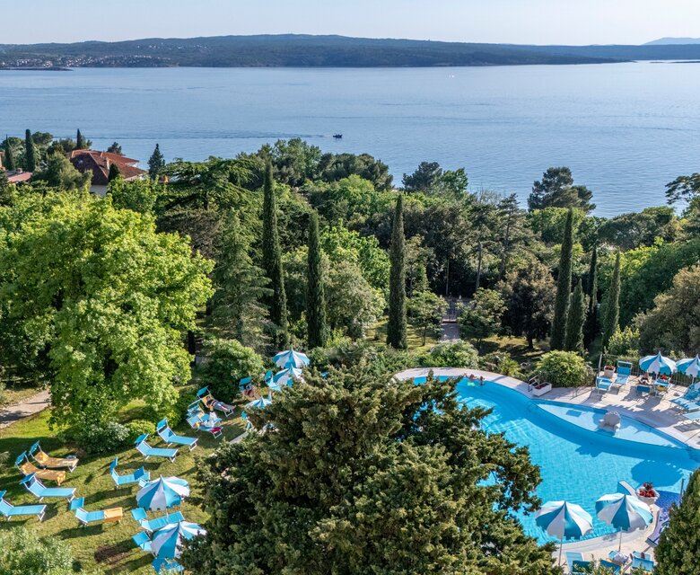 Blick auf einen Pool umgeben von üppigem Grün und Liegen mit Sonnenschirmen. Im Hintergrund erstreckt sich der ruhige See und die sanften Hügel der Umgebung. Einladende Atmosphäre für Entspannung und Erholung.