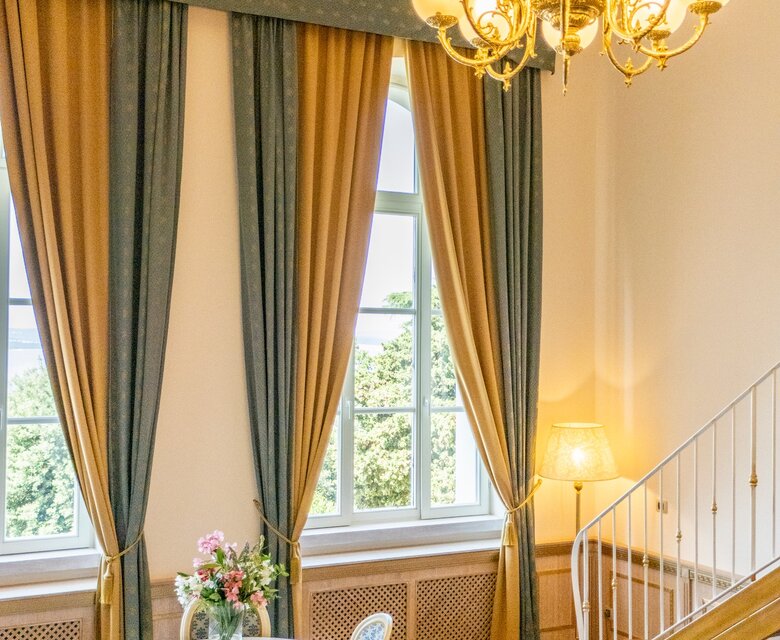 Heller, eleganter Raum mit großen Fenstern, die viel Tageslicht hereinlassen. Ein runder Tisch mit vier Stühlen steht in der Mitte, umgeben von stilvollen Vorhängen. Eine gemütliche Couch und ein Kronleuchter verleihen dem Raum eine einladende Atmosphäre.