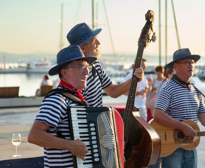Drei Musiker in gestreiften Hemden spielen Instrumente am Hafen. Im Hintergrund sind Yachten und ein sanfter Sonnenuntergang zu sehen. Einige Gläser stehen auf einem Tisch in der Nähe. Die Atmosphäre ist festlich und entspannt.