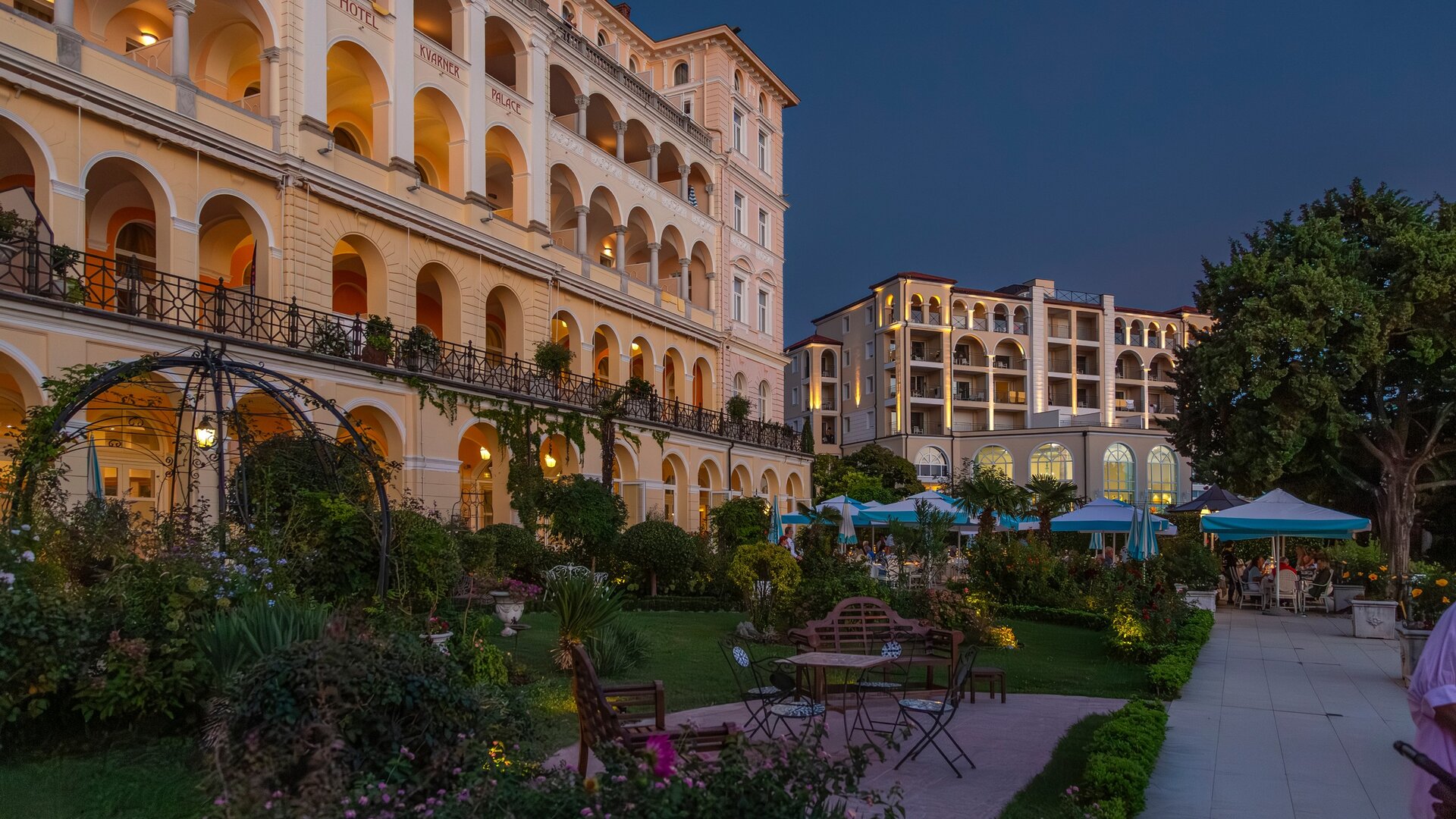 Eine elegante Hotelanlage mit mehreren Etagen, umgeben von einem gepflegten Garten. Die Architektur ist klassisch mit Bögen und Balkonen. Bei Dämmerung beleuchtet, schaffen die Lichter eine einladende Atmosphäre. Gäste genießen die Außenbereiche unter Sonnenschirmen.