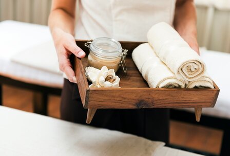 Eine Person hält ein Holztablett mit zwei gerollten Handtüchern, einem Glas mit einer Creme und einer dekorativen Blume. Im Hintergrund sind Massagetische zu sehen, die eine entspannende Atmosphäre... Eine Person hält ein Holztablett mit zwei gerollten Handtüchern, einem Glas mit einer Creme und einer dekorativen Blume. Im Hintergrund sind Massagetische zu sehen, die eine entspannende Atmosphäre schaffen.