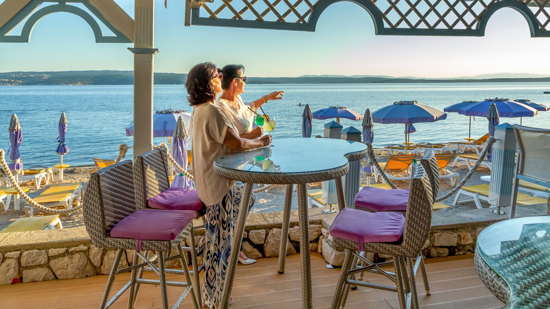 Zwei Frauen sitzen an einem Tisch mit Blick auf das Wasser, während sie einen Drink genießen. Im Hintergrund sind Liegen und Sonnenschirme am Strand zu sehen, und die ruhige Wasseroberfläche reflektiert das Licht der Abendsonne.