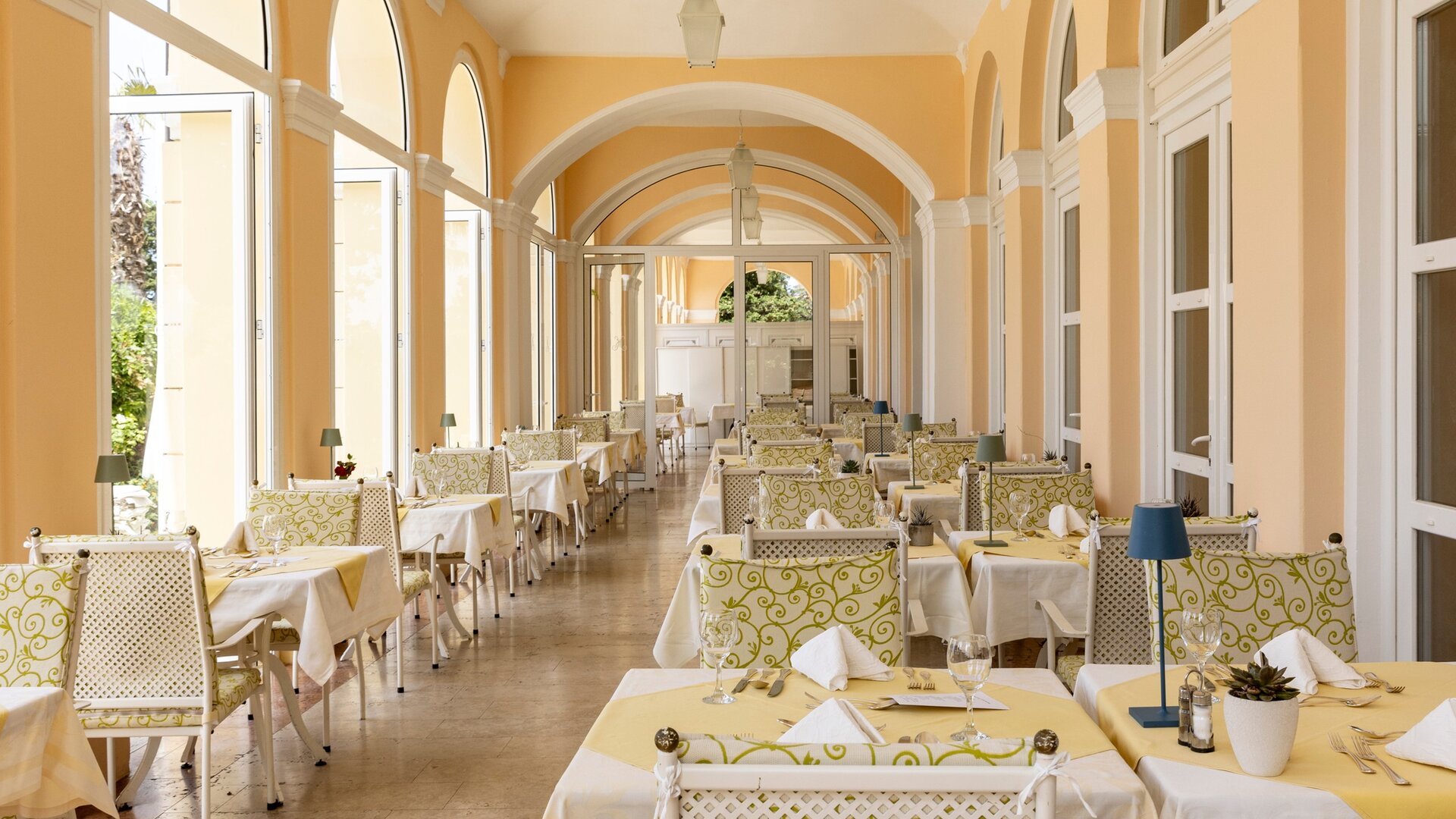 Helles Restaurant mit eleganten Tischen und Stühlen, umgeben von großen Fenstern, die viel Tageslicht hereinlassen. Die Wände sind in sanften Gelbtönen gehalten, und die Tische sind mit weißen Tischdecken und dekorativen Elementen gedeckt.