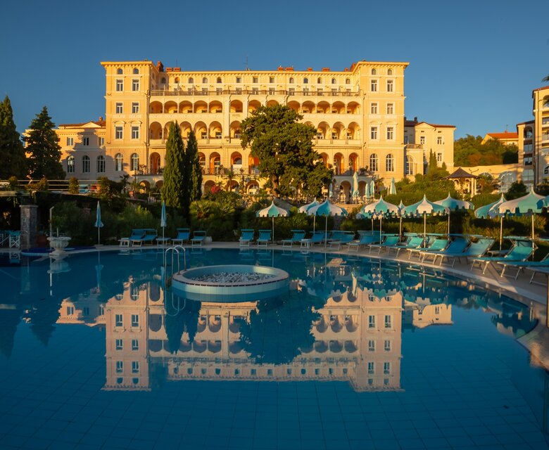 Ein elegantes Hotelgebäude spiegelt sich im klaren Wasser eines Pools, umgeben von grünen Bäumen und Liegen. Die Abendsonne taucht die Szene in warmes Licht und schafft eine entspannte Atmosphäre.