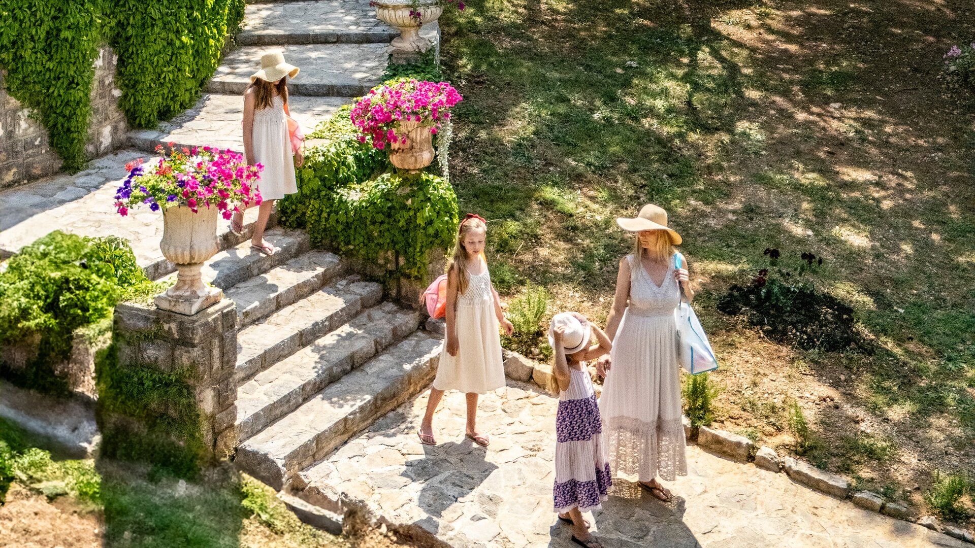 Eine Frau mit einem Sonnenhut steht auf einer Steintreppe und hält die Hand eines Mädchens. Zwei weitere Mädchen in hellen Kleidern spielen im Hintergrund. Bunte Blumen in Töpfen schmücken die Treppe, umgeben von grünem Gras und Bäumen.