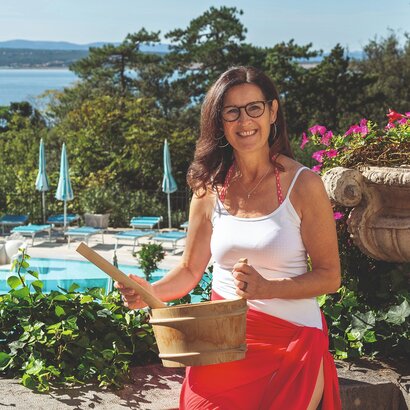Eine Frau mit langen, lockigen Haaren und einer roten Badehose steht am Pool. Sie hält einen Holzbehälter in der Hand und lächelt. Im Hintergrund sind Sonnenliegen und ein schöner Blick auf das Wasser und die Landschaft zu sehen.