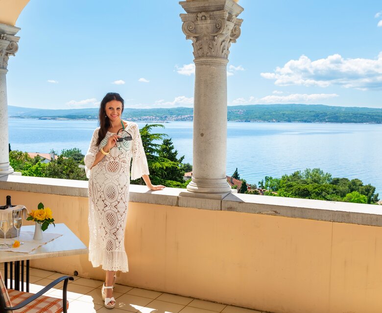 Eine Frau in einem weißen, luftigen Kleid steht auf einem Balkon mit Blick auf das glitzernde Wasser und die umliegende Landschaft. Auf dem Tisch neben ihr stehen eine Flasche Sekt und ein Glas. Die Atmosphäre ist entspannt und sonnig.