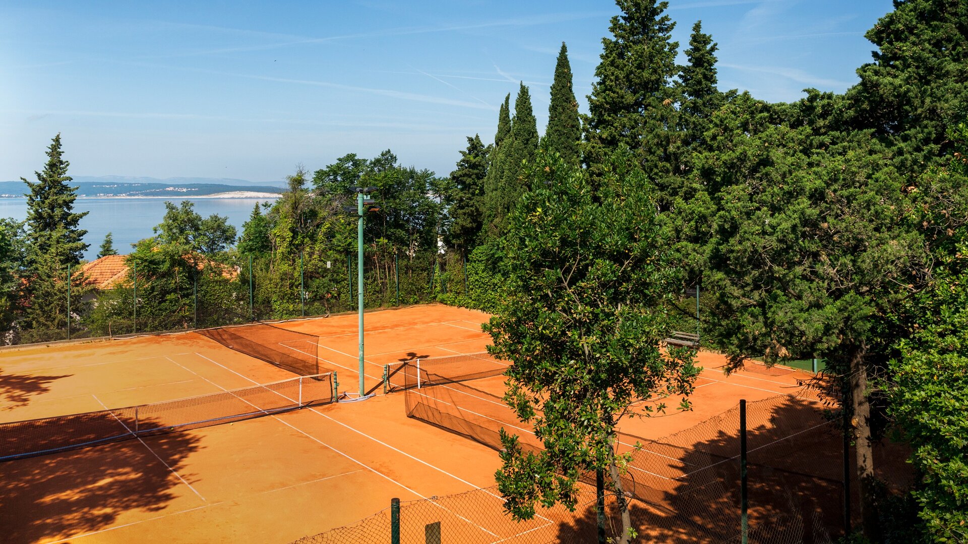 Tennisplätze mit rotem Sand, umgeben von üppigem Grün und hohen Bäumen. Im Hintergrund ist ein Blick auf das Wasser und den klaren Himmel zu sehen, was eine entspannte und einladende Atmosphäre schafft.